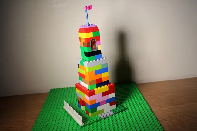 Colorful Lego skyscraper built on a green baseplate with a small pink flag on top, showcasing a childhood craft project.