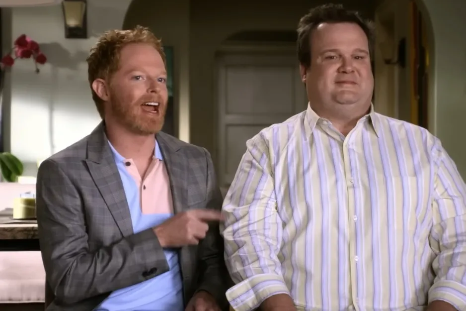 Two men from Modern Family talking indoors, one in a gray blazer and the other in a striped shirt sitting together.