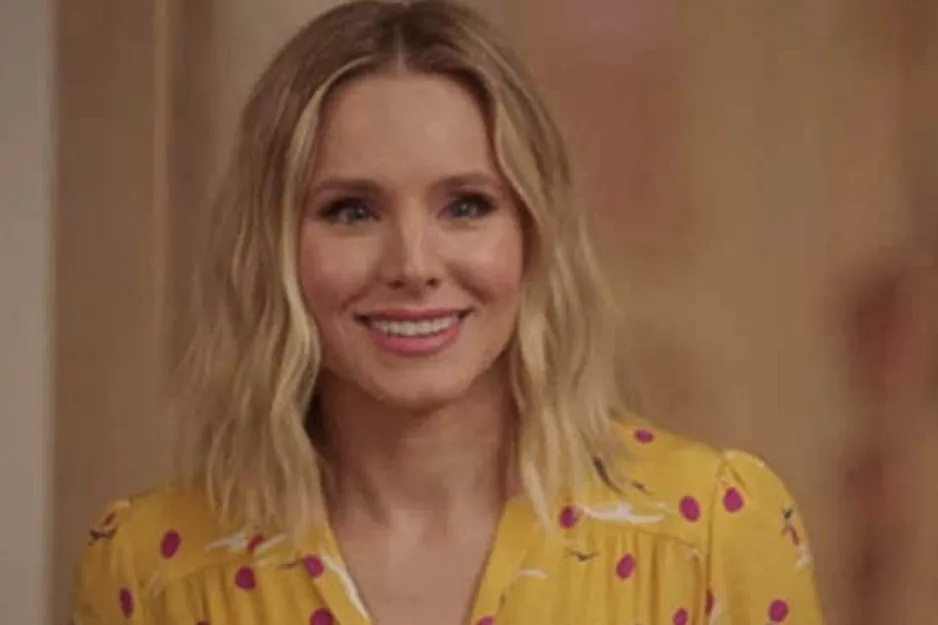 Smiling woman with shoulder-length blonde hair wearing a yellow dress with red and white patterns in a warm setting.