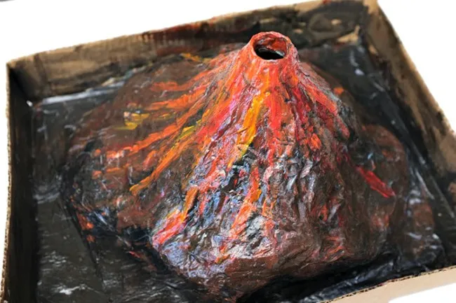 Science fair volcano project painted with red and orange lava details inside a black cardboard tray for a childhood craft.