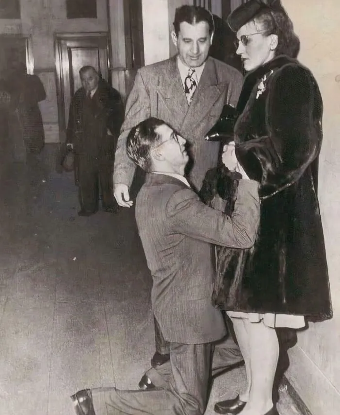 Black and white historical photo showing a man kneeling in front of a woman wearing a hat and coat indoors.