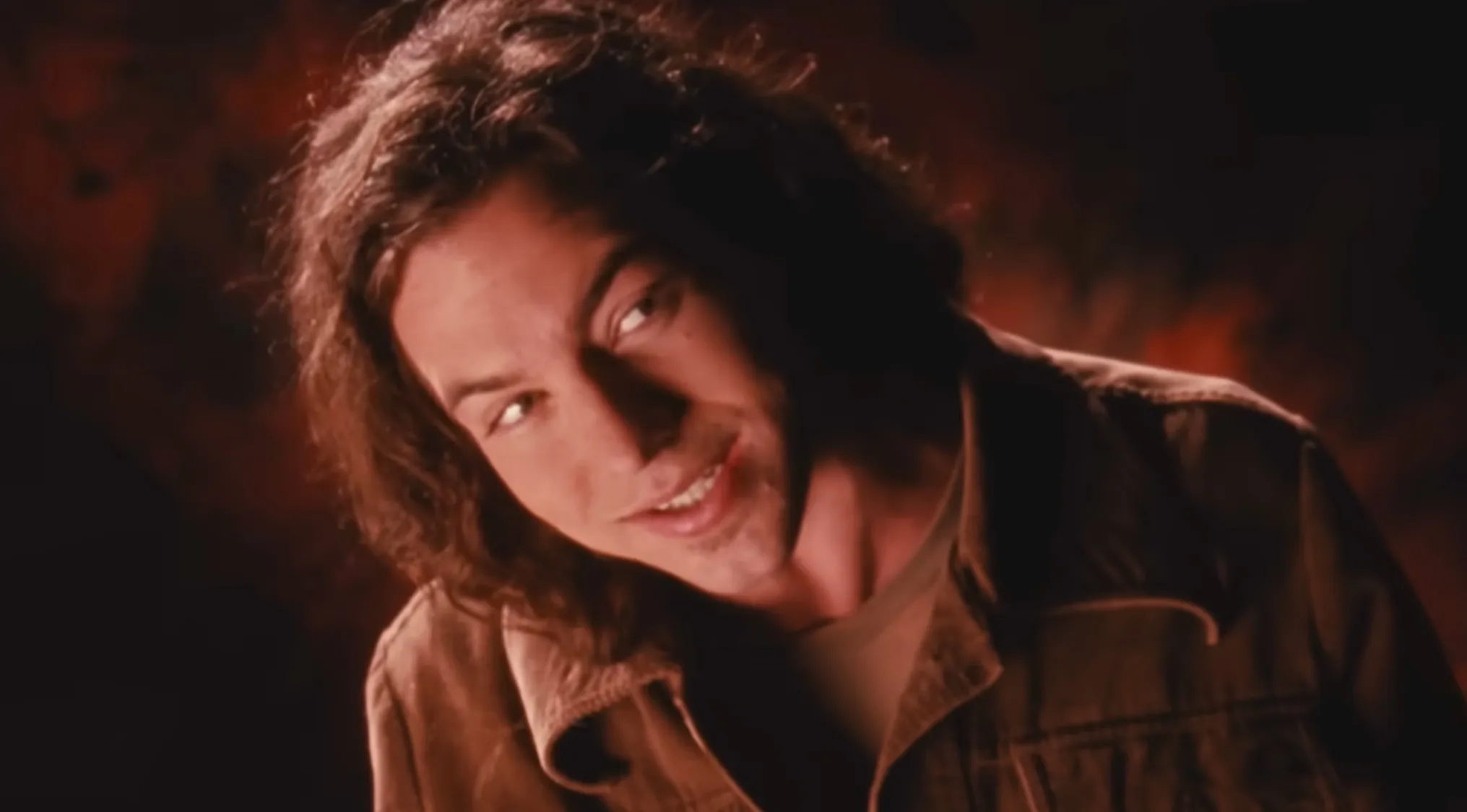 Eddie Vedder, lead singer of Pearl Jam, performing in a dimly lit setting with long hair and casual jacket.