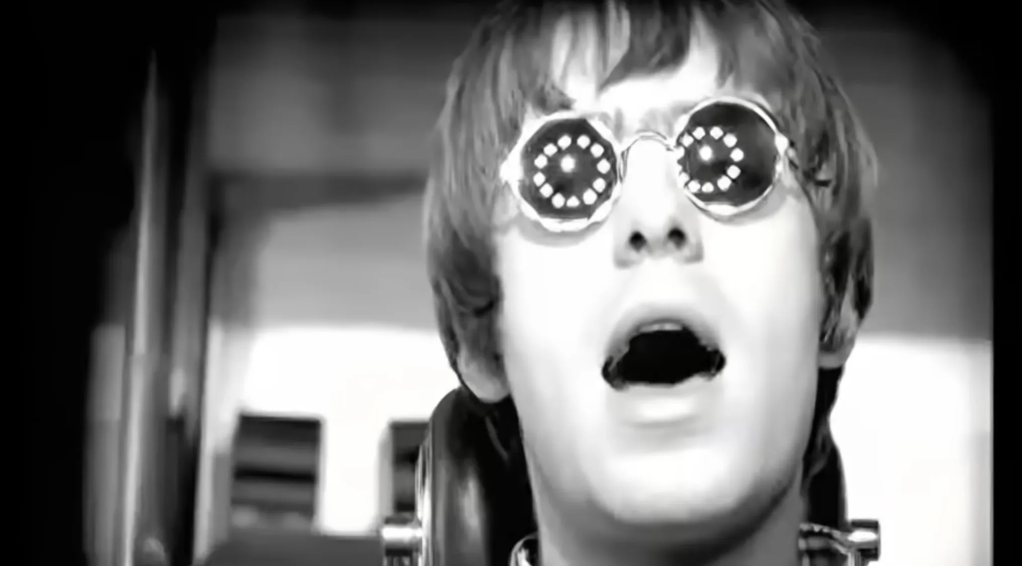 Liam Gallagher wearing round glasses with lights, singing as the frontman of a famous band in a black and white scene.