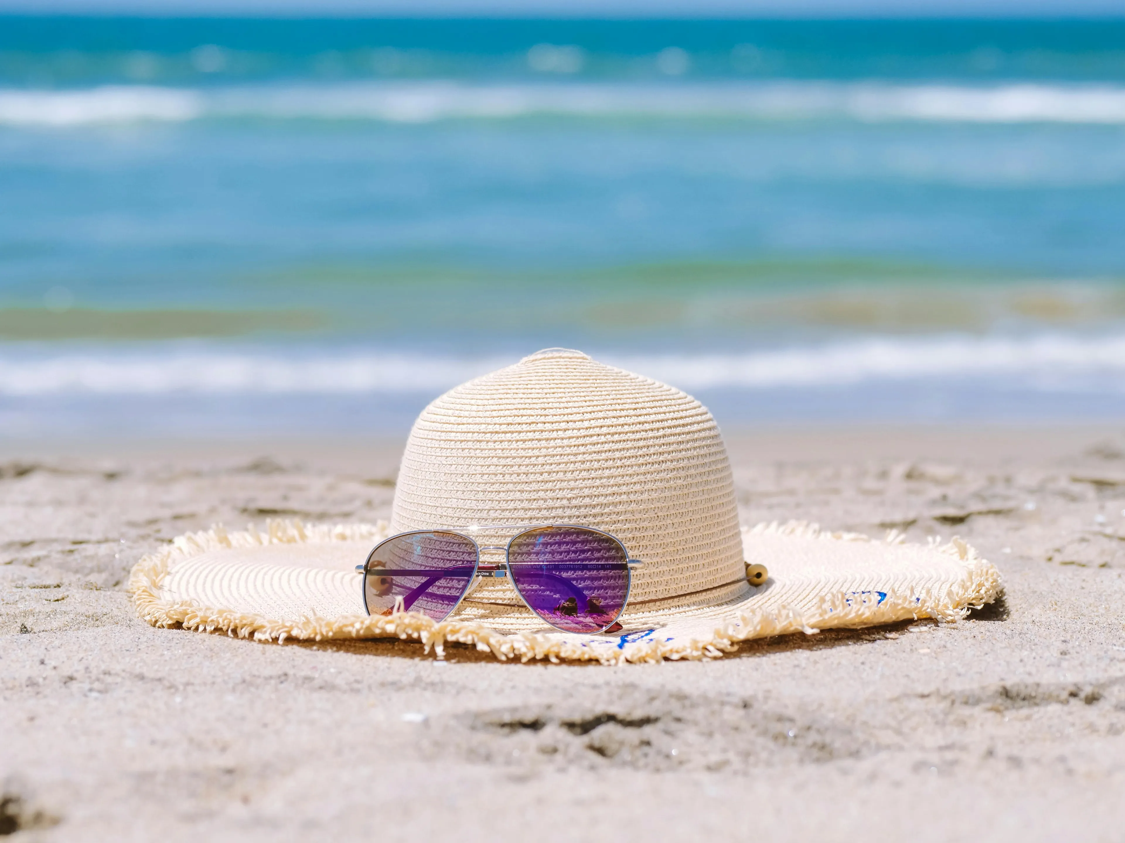 Straw summer hat and sunglasses resting on sandy beach with ocean waves in the background on a sunny day.