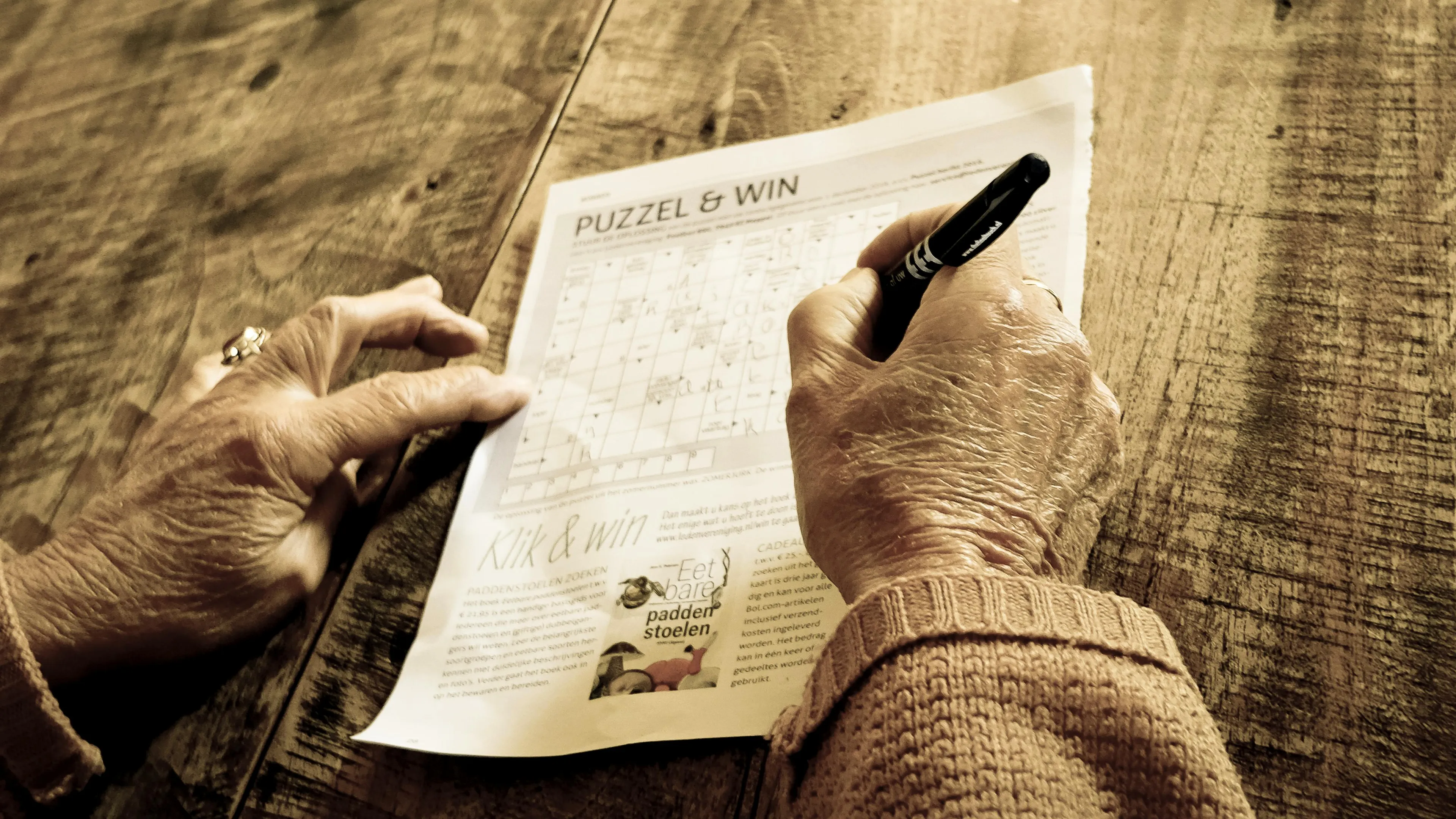 Person solving a crossword puzzle in a newspaper at a wooden table, enjoying a quiet indoor activity.