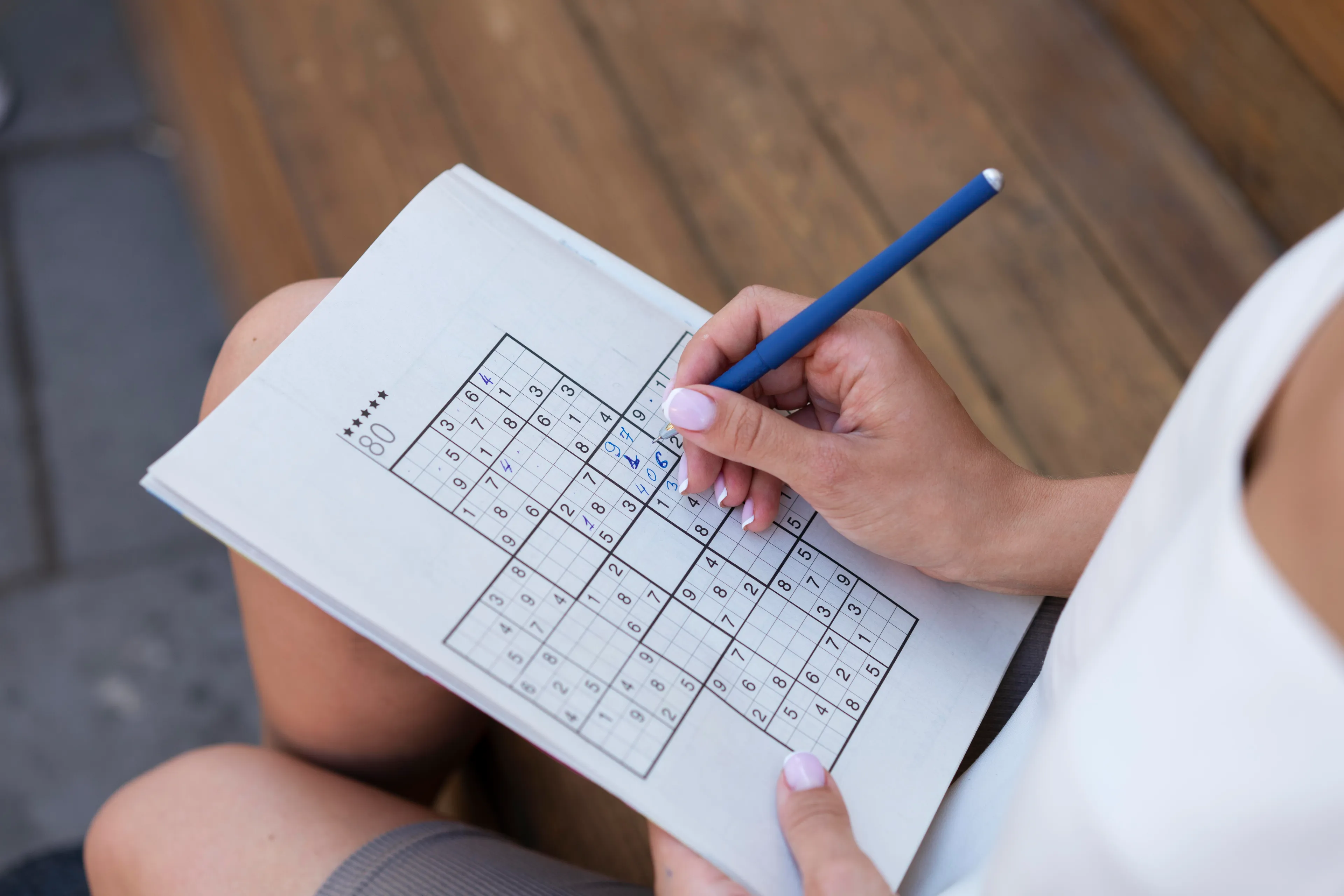 Person solving a Sudoku puzzle with a pencil on paper, focusing on numbers and logic in a relaxed setting.