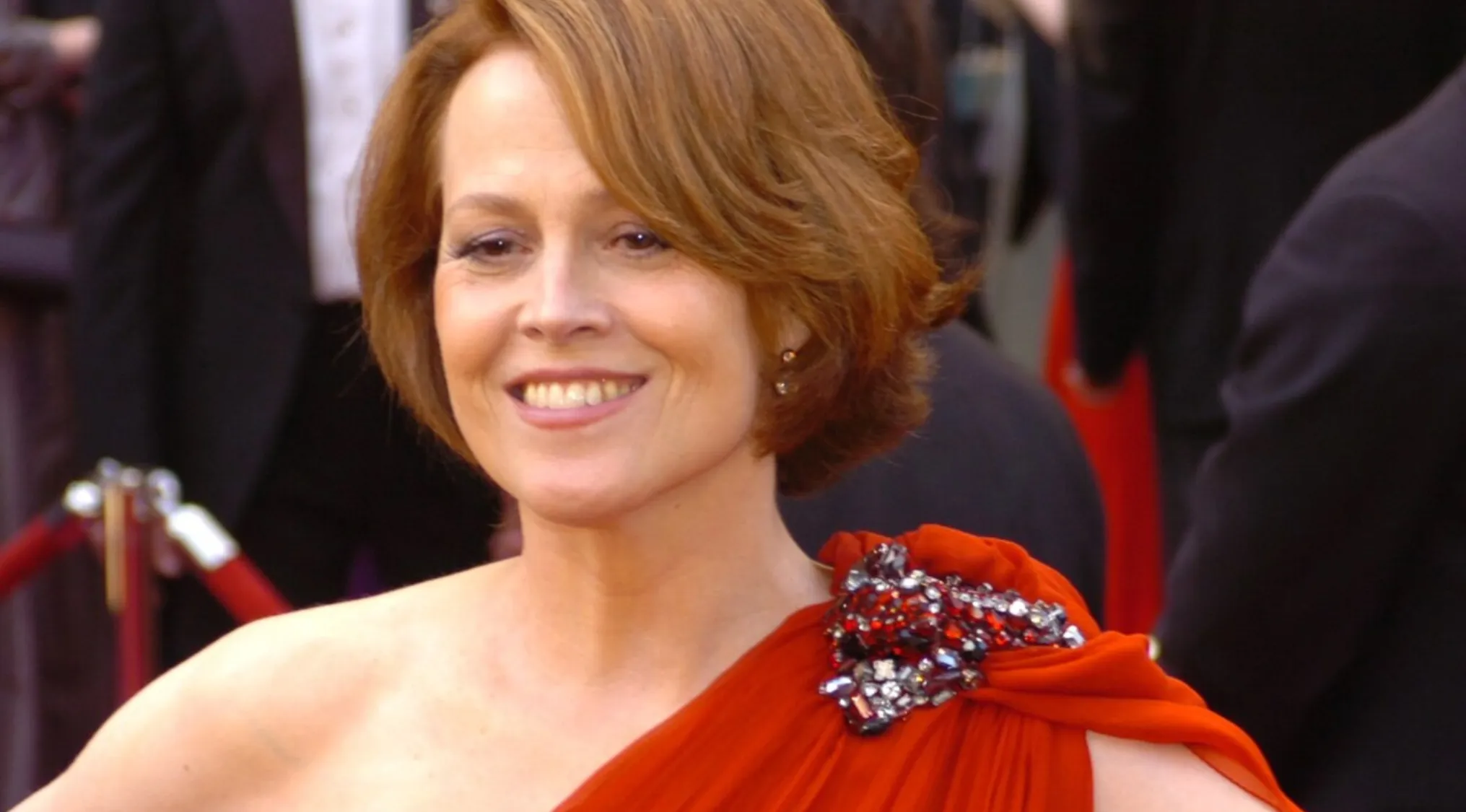 Sigourney Weaver smiling on red carpet wearing red dress with jeweled detail guess the movie cast Alien survivor