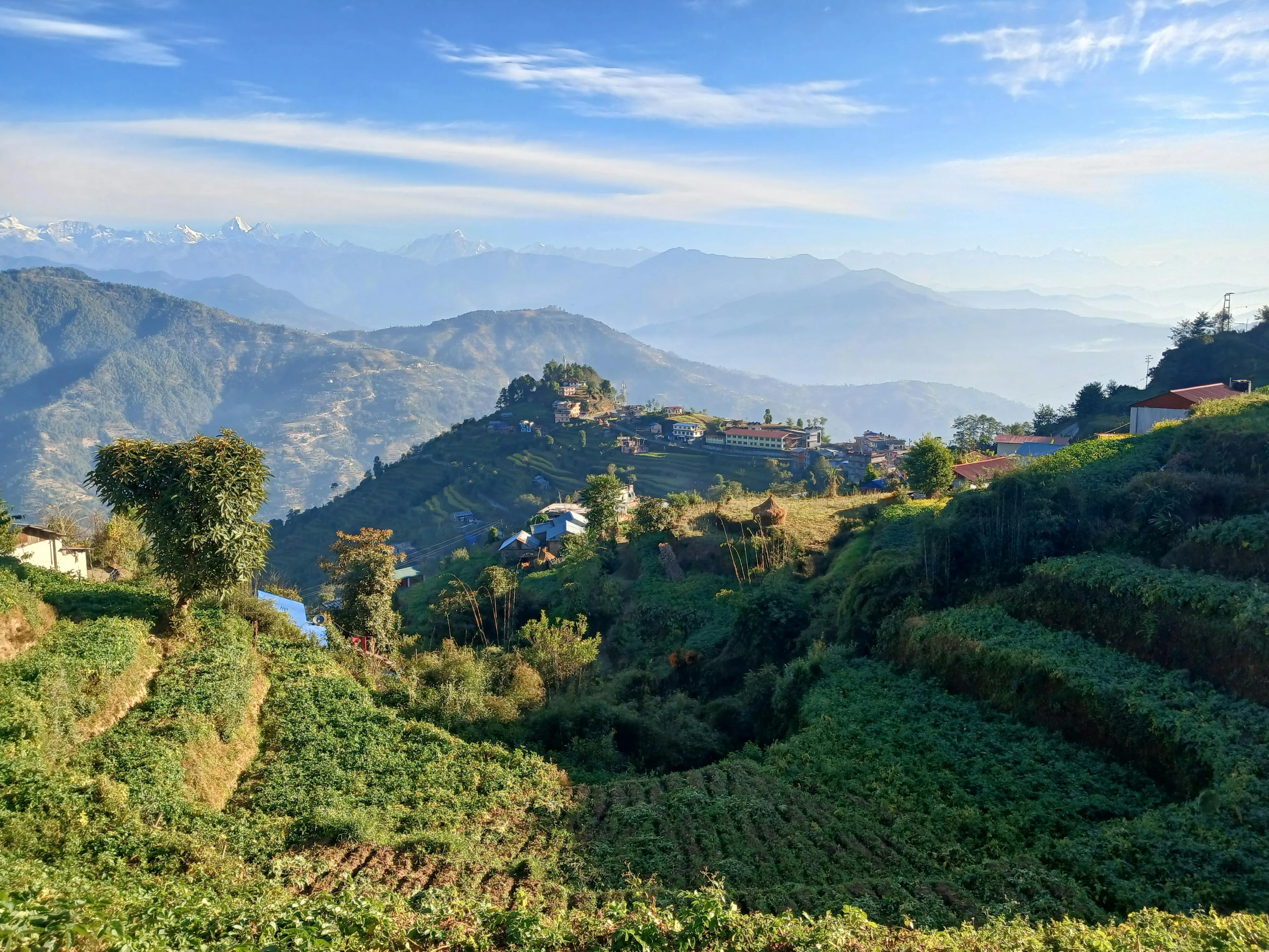 Hilly landscape with terraced fields and distant mountains in Nepal under a clear blue sky.