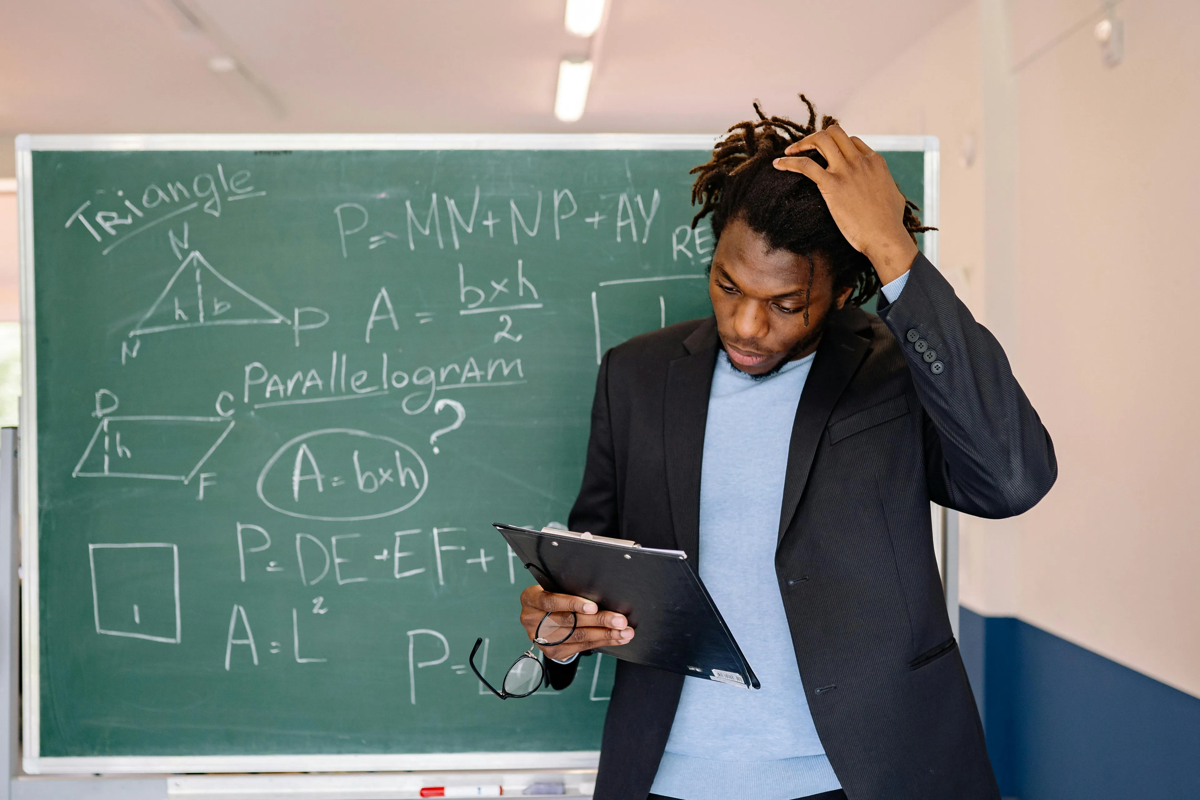 Man in blazer scratching head while holding clipboard in front of blackboard with difficult math and quiz questions about numbers