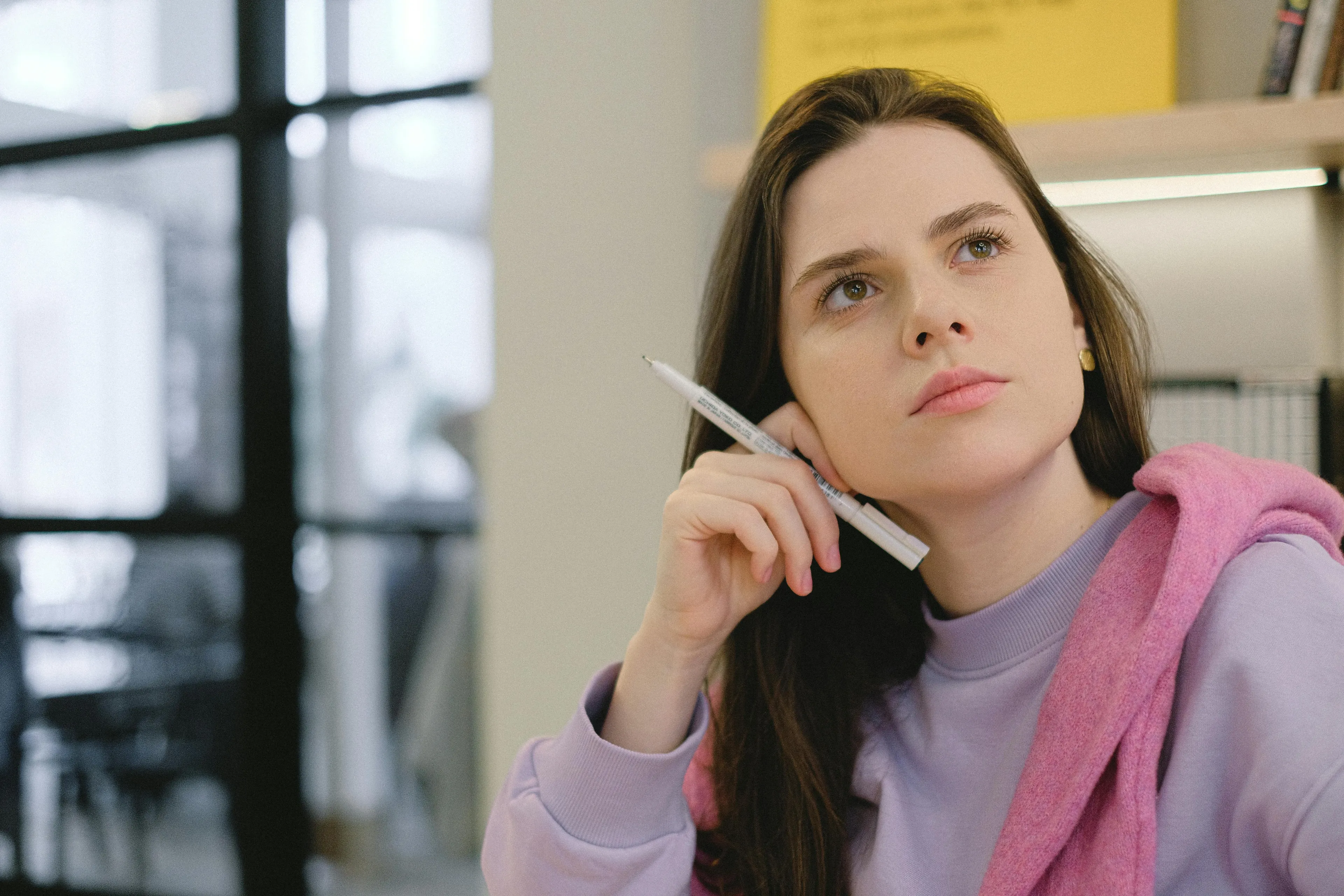 Young woman in a purple sweater and pink scarf holding a pen, deep in thought during a difficult general knowledge quiz.