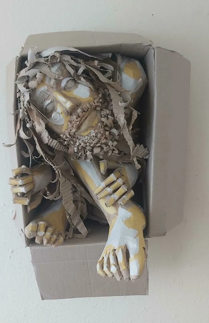 A sculpture of a figure made from cardboard and paper, painted yellow and white, emerging from a box. Trash art.