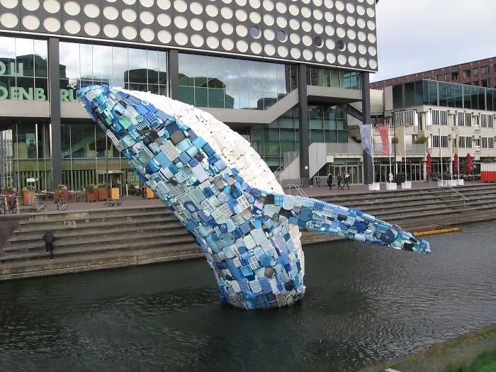 A large whale sculpture made from recycled blue and white trash rising from water, a creative way to use trash.