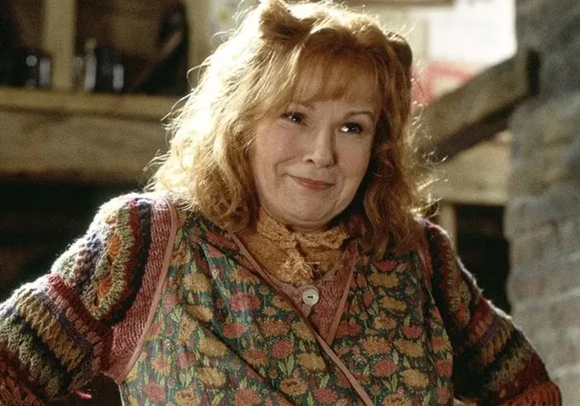 Mrs. Weasley smiling warmly, wearing a colorful knitted sweater and floral vest in a cozy rustic setting.