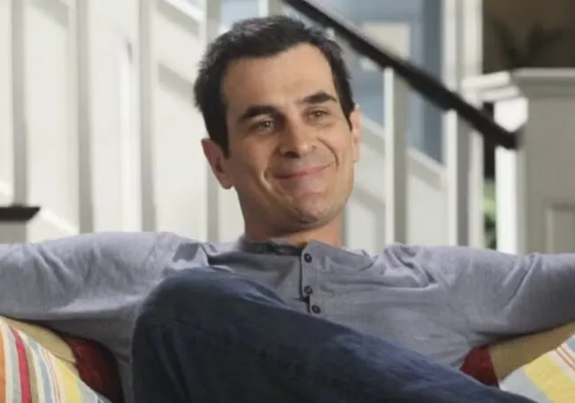 Phil Dunphy smiling relaxed on a couch wearing a casual grey shirt in a bright home setting.