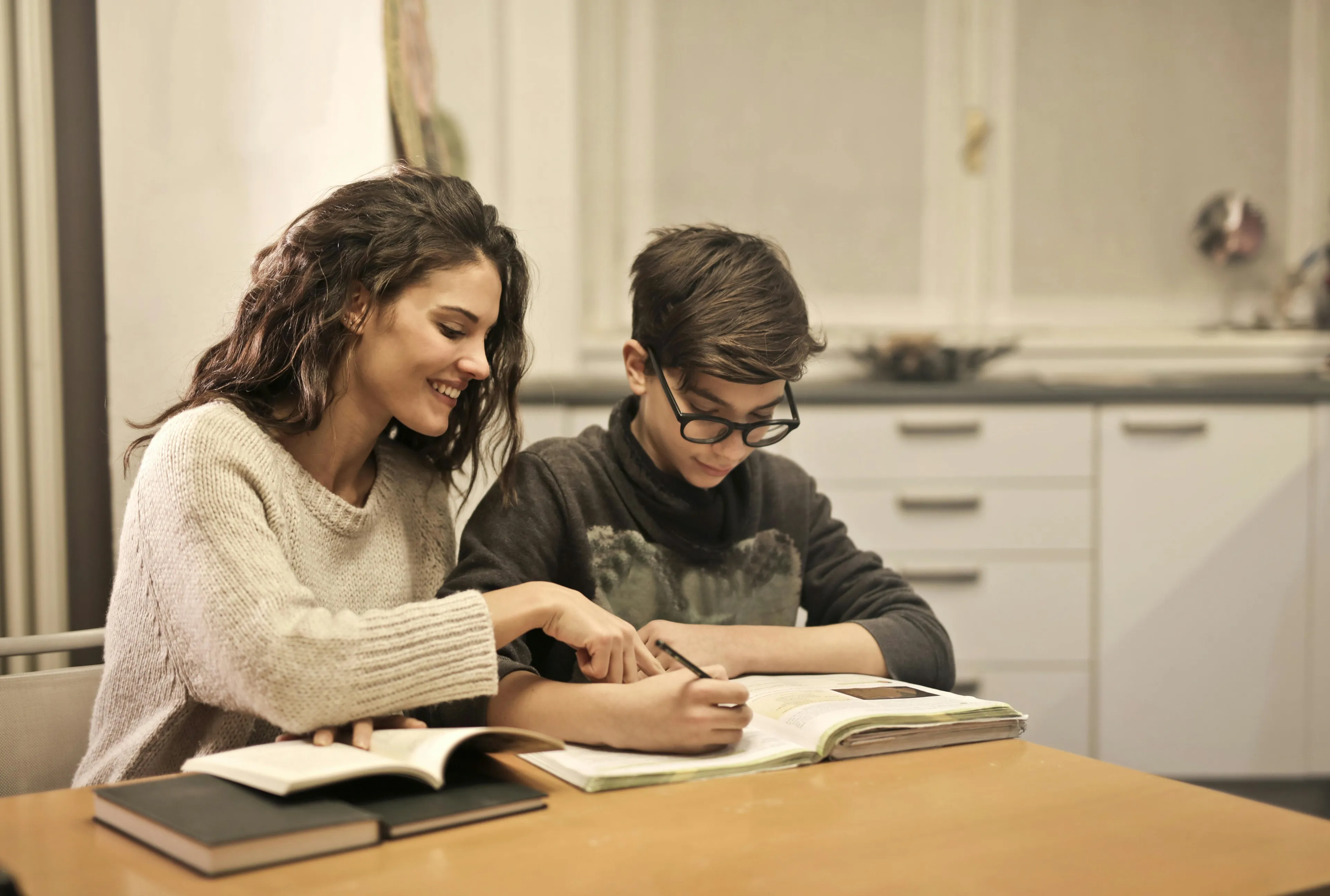 Mother helps her son with homework hour, both smiling and focused on the assignment at the kitchen table.