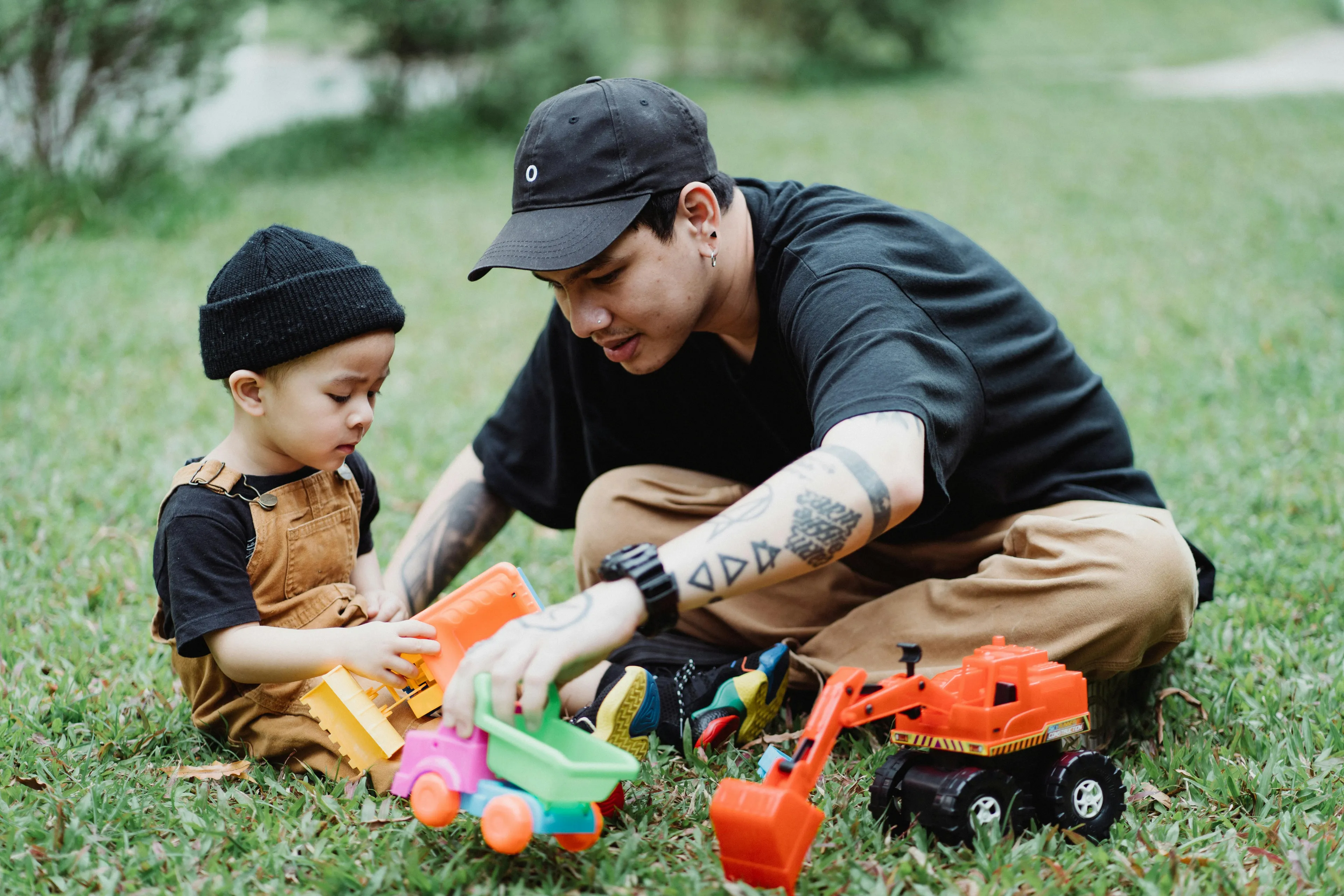 Father and toddler playing outdoors on grass with colorful toy trucks, enjoying outdoor play and quality time together.