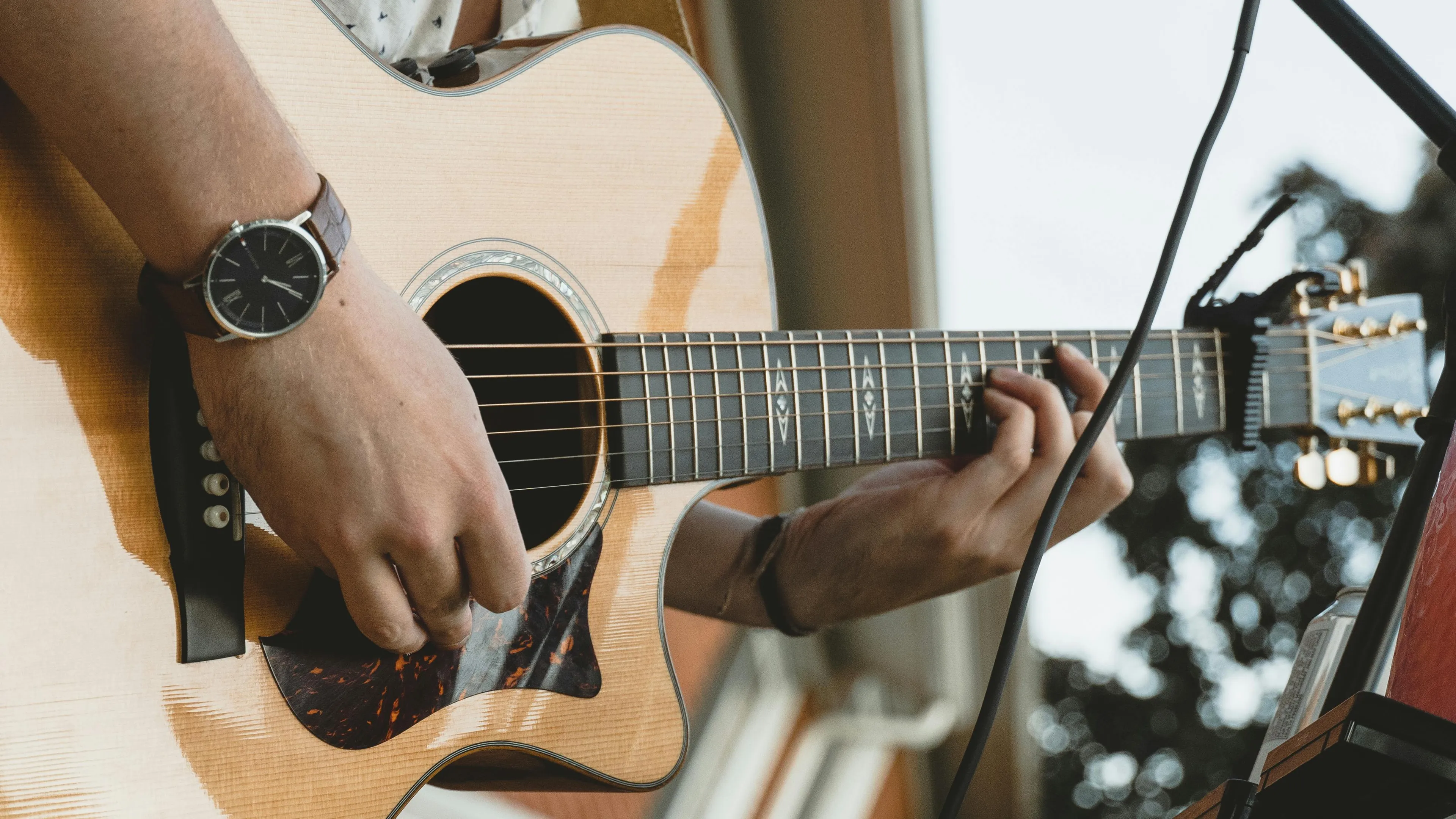 Close-up of hands playing an acoustic guitar, showcasing detailed fretboard and natural wood finish in a relaxed setting.