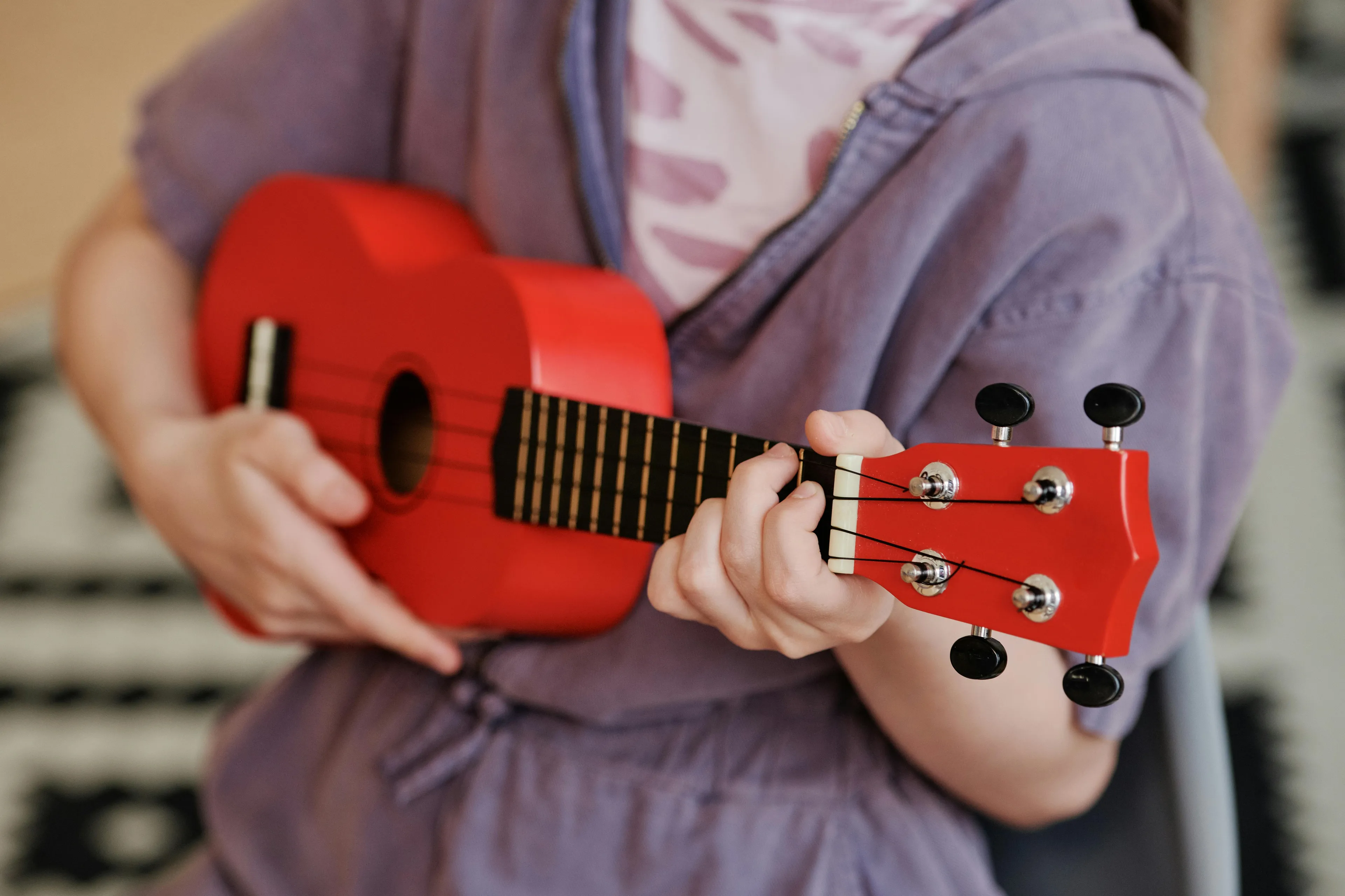 Person wearing a casual shirt playing a red ukulele, focusing on the strings and hand positioning on the instrument's neck.