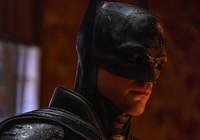 Batman in a dark leather suit with a wet texture, looking serious and focused in a dimly lit setting.