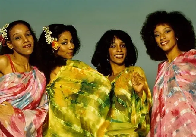 Four women in colorful flowing dresses smiling together representing family and parenting playlist anthem vibes.