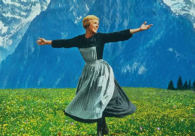 A joyful woman in traditional dress dancing in a meadow with mountains in the background from The Sound of Music.
