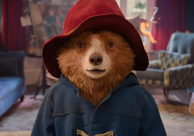 Paddington bear wearing a red hat and blue coat sitting in a cozy living room, family movie choice inspiration.
