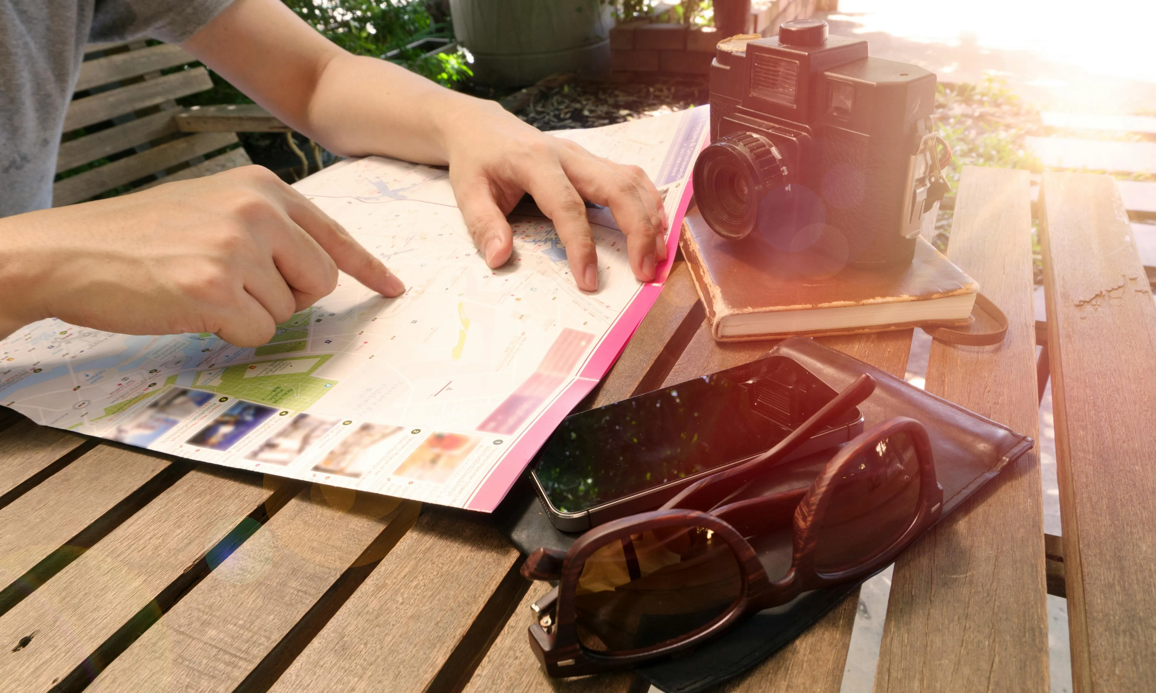Person planning a vacation using a map on a wooden table with sunglasses, phone, and vintage camera nearby.
