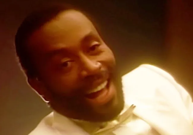 A smiling man wearing a white suit and bow tie in a warm, softly lit setting, representing parenting playlist anthem.