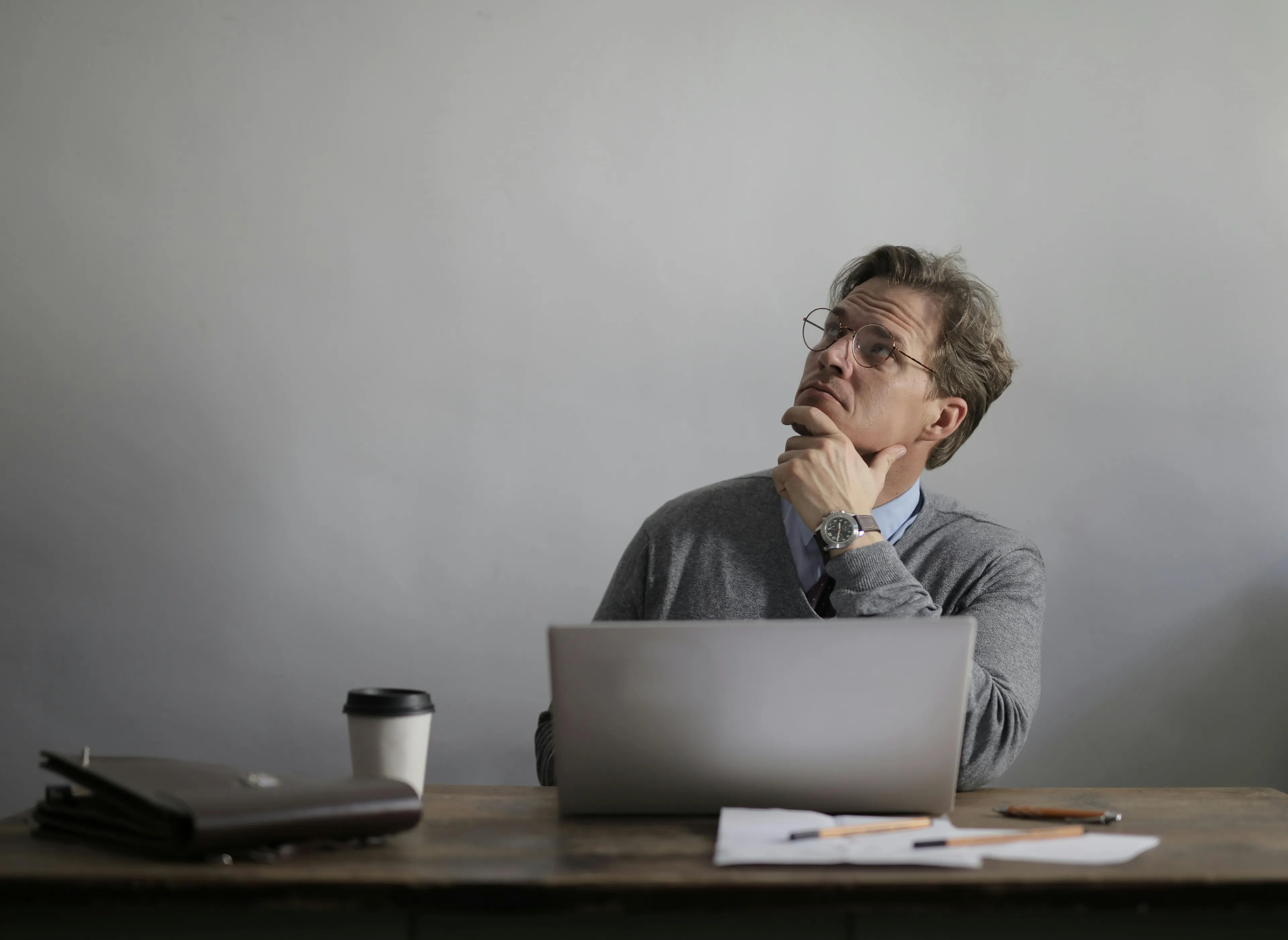 Man in glasses sitting at a desk with laptop, coffee, and documents, thinking about geography connections and places.