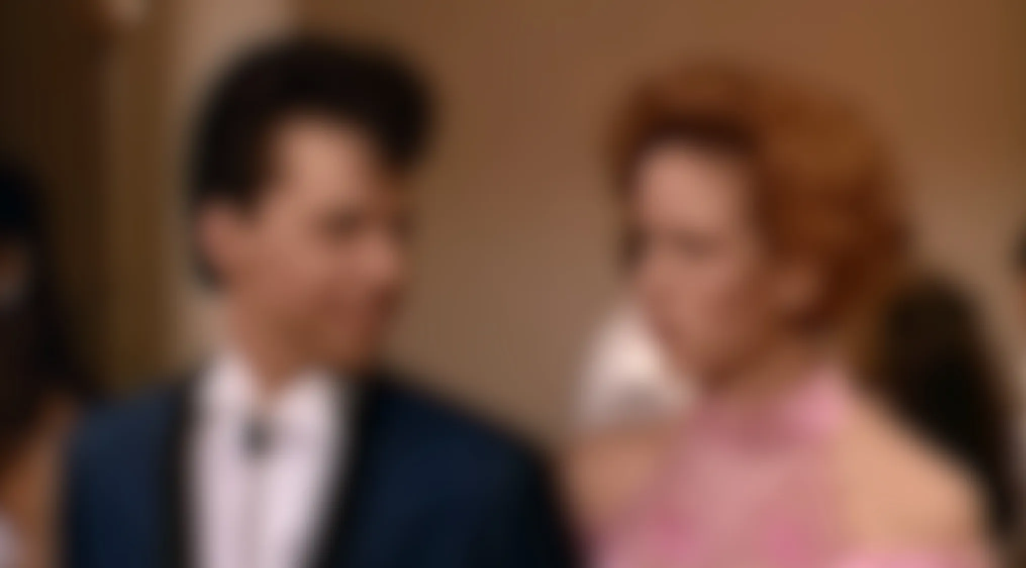 Blurry movie scene of a man in a dark suit and high hair looking towards a woman with curly reddish hair in a pink dress.