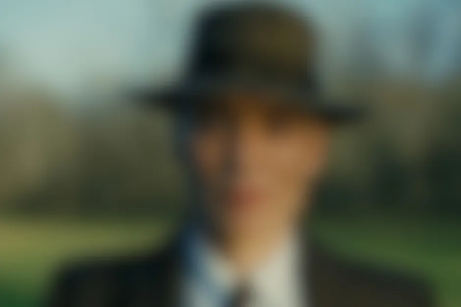 Blurred scene from Oppenheimer showing a character in a hat and suit, outdoors with a light background.