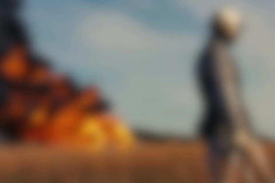 Saving Private Ryan scene: A blurred figure stands in a field, observing a massive fire and smoke under a pale sky.