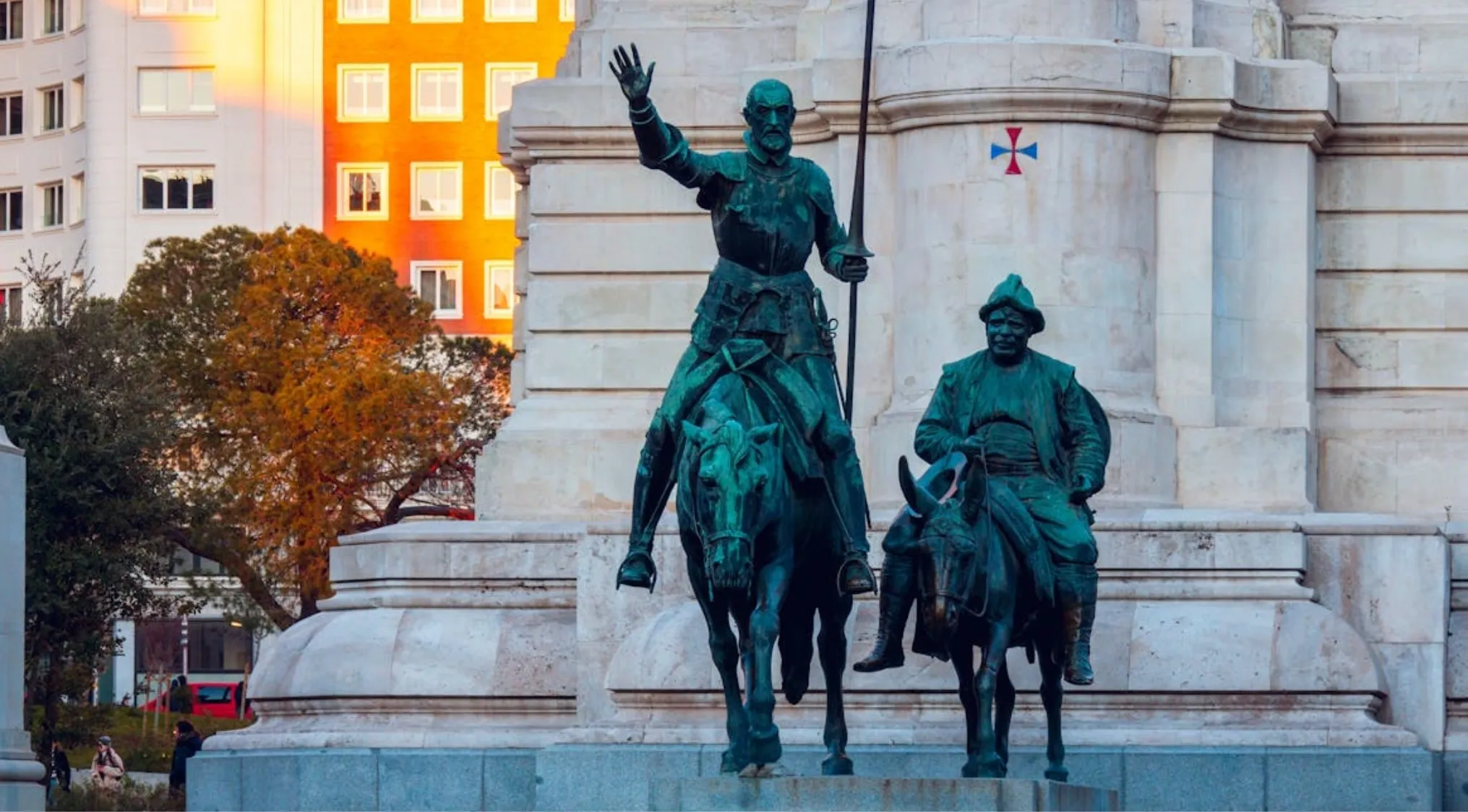 Bronze statues of Don Quixote on horseback with arm raised and Sancho Panza on his donkey, in front of a monument.