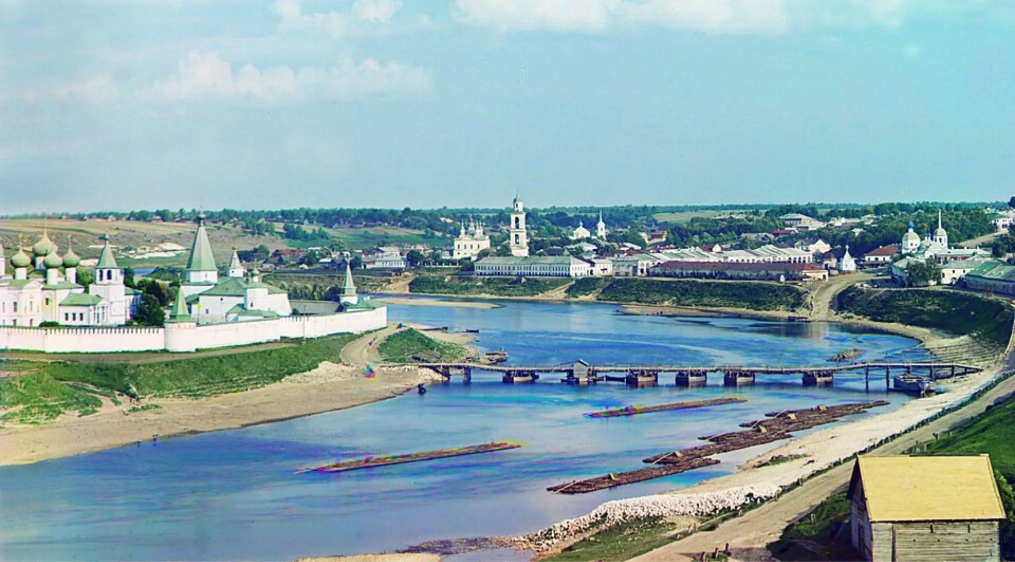 View of a historic town and monastery by the River Volga, with a wooden bridge and log rafts. Scenic Russian landscape.