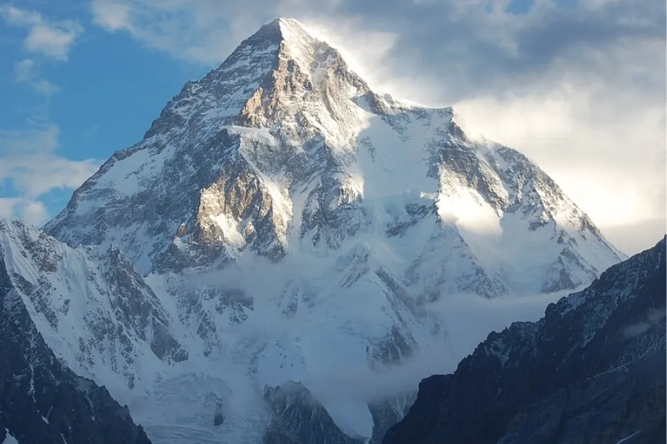 K2 mountain, snow-covered and majestic, partially hidden by clouds with sunlit peaks and rugged terrain.