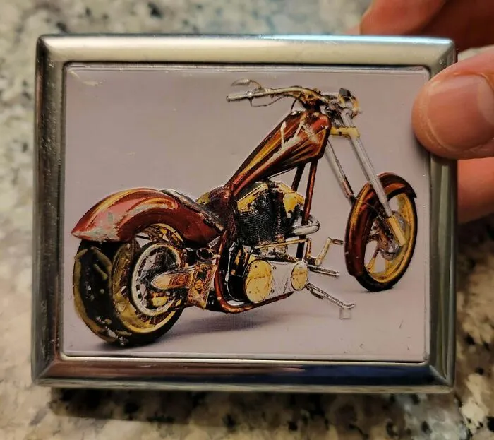 A hand holds a gold bar-shaped lighter case with a custom motorcycle design, a unique Goodwill bin find.