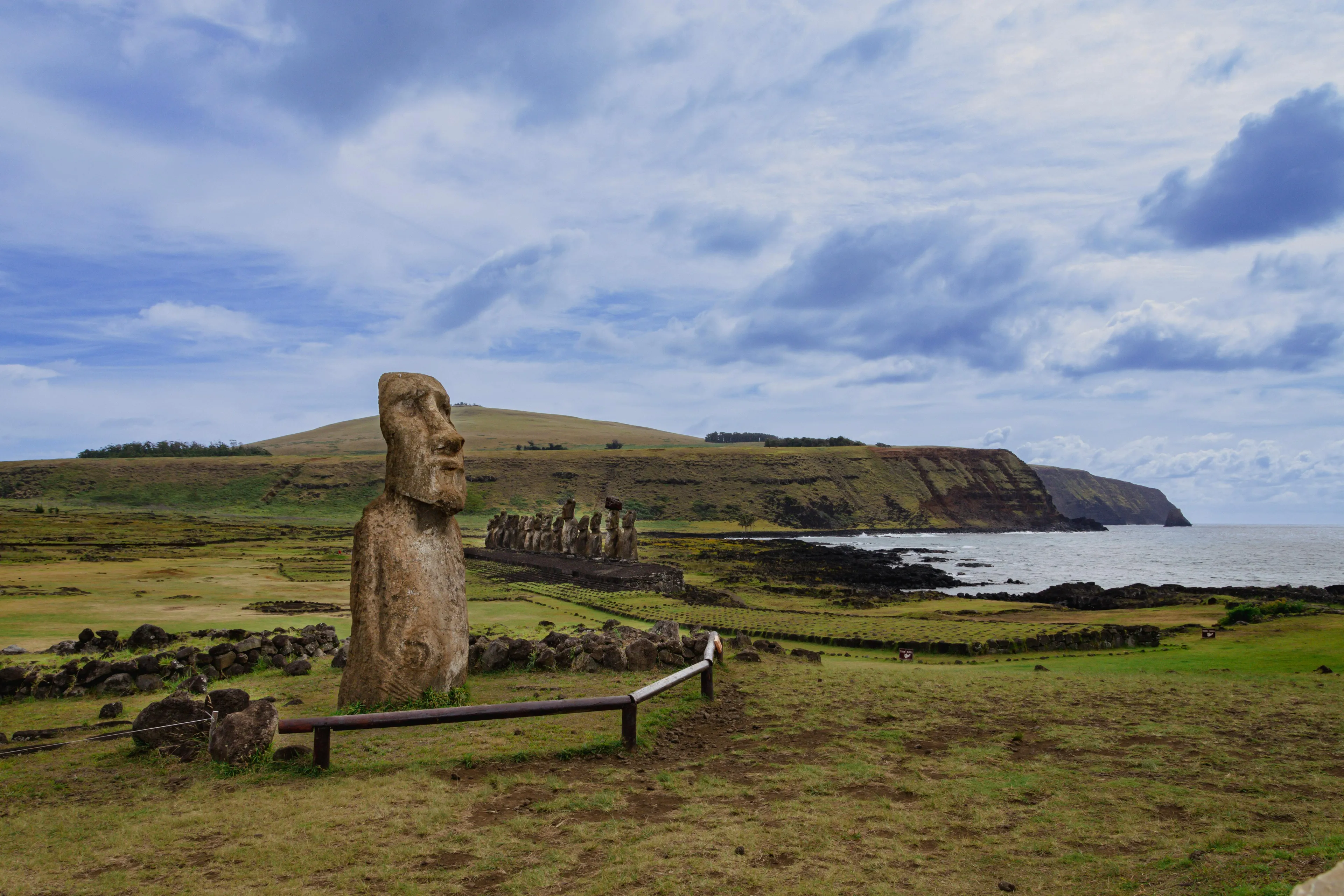 Massive Moai statue on Easter Island, with more in the distance, alongside coastline. Guess the country from this landmark.