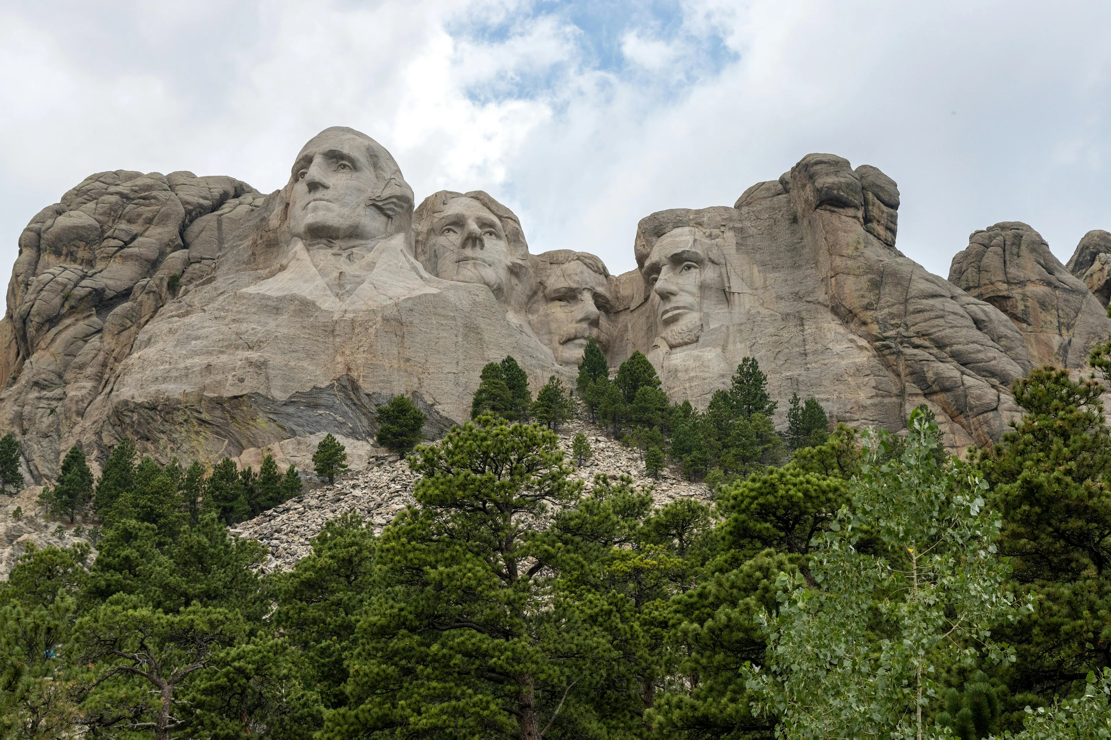 MOUNT RUSHMORE, featuring four carved US presidential faces on a rocky mountain, with green pine trees in the foreground.