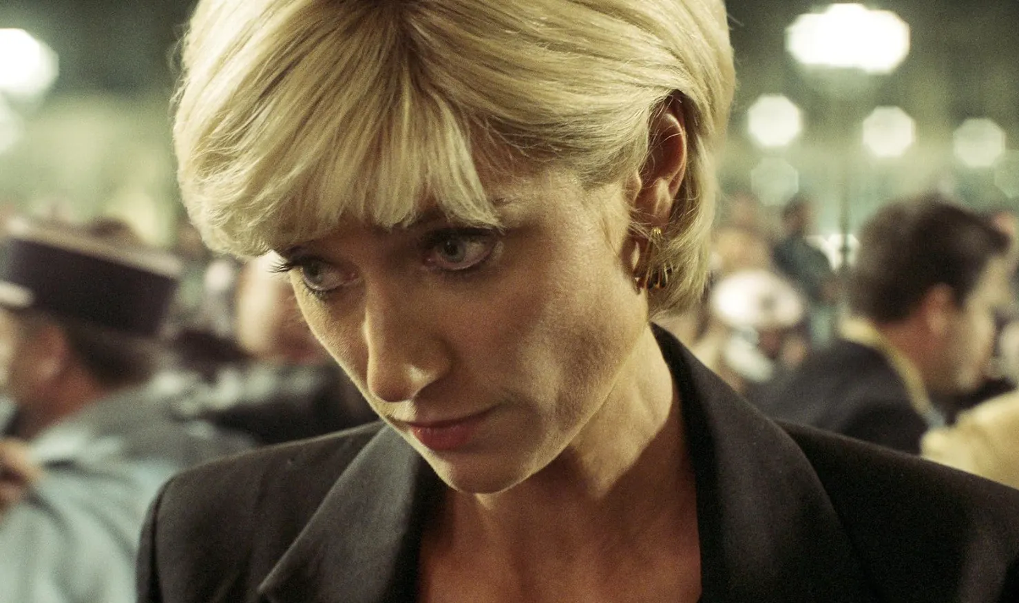 A close-up of a woman in The Crown with short blonde hair in a crowded, dimly lit room.