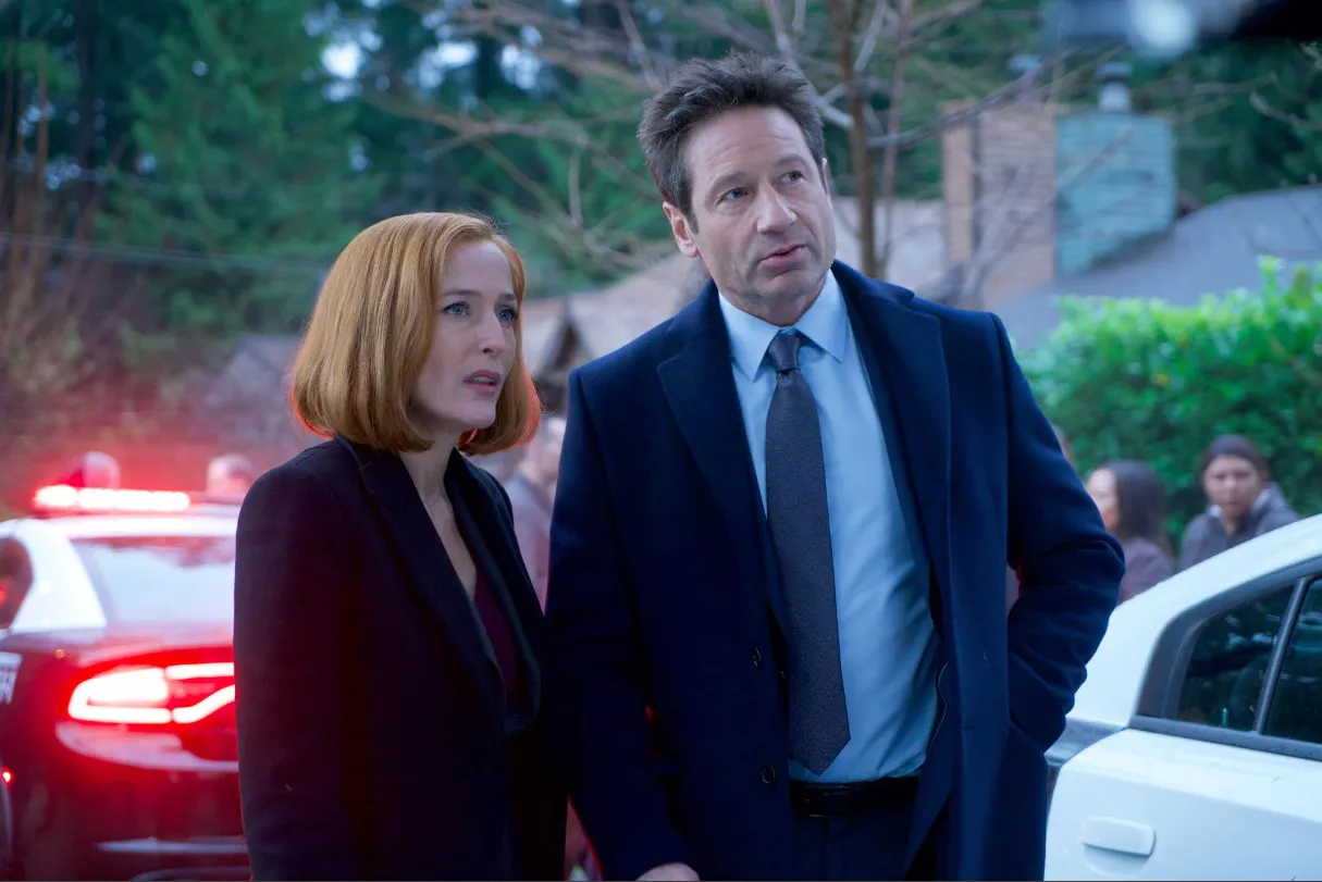 Two detectives from The X-Files discussing a case outdoors near police cars with flashing lights.