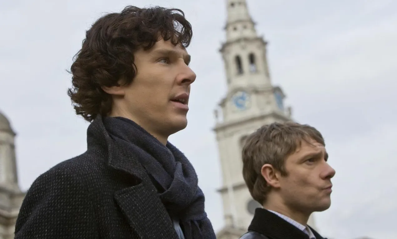Actors from Sherlock standing outside with a church tower in the background, dressed in dark coats and scarves.