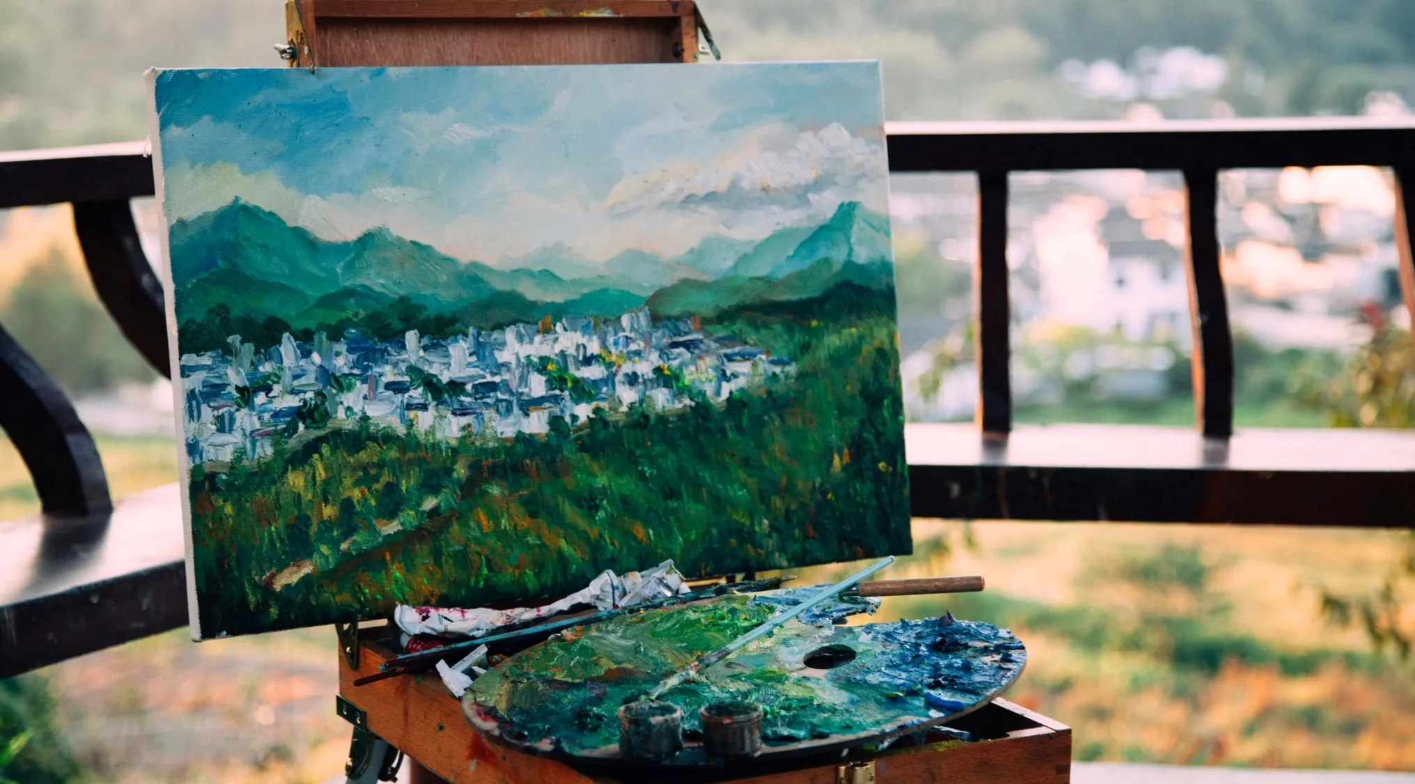 Landscape painting on an easel showing the area closest to the viewer with green trees and town details in focus.