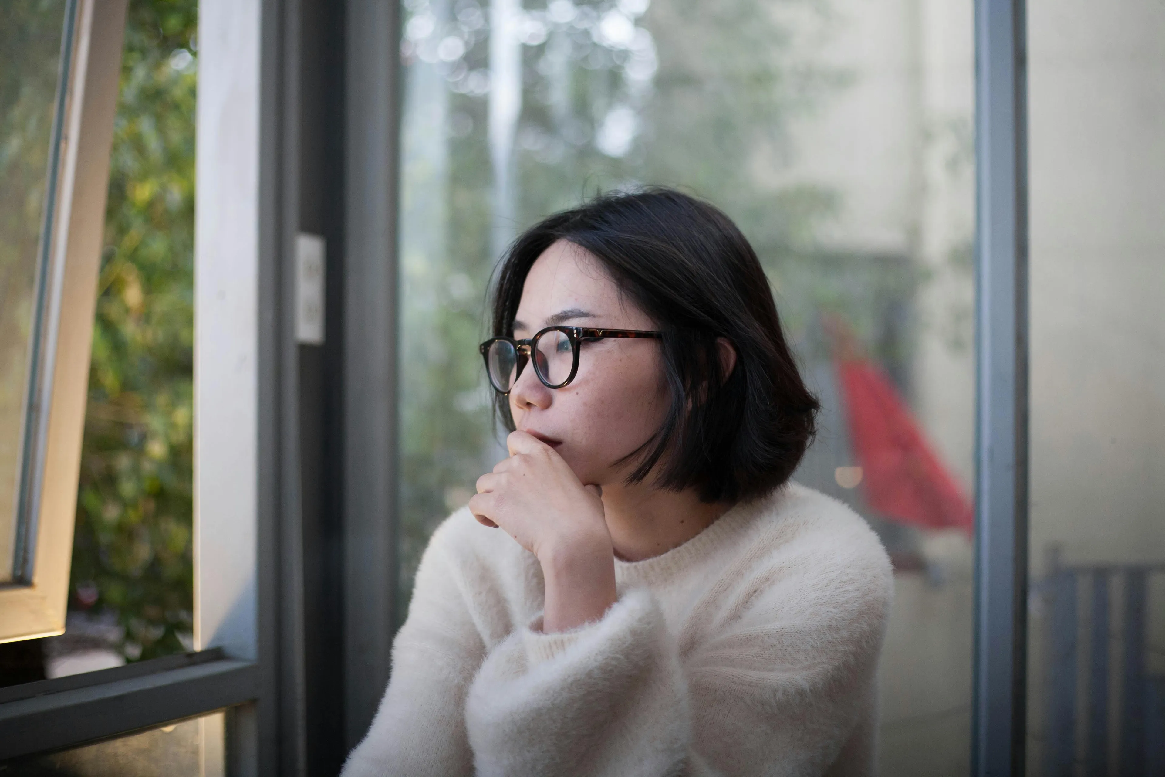 Young woman wearing glasses and a white sweater looking thoughtfully out a window, representing human body biology study.