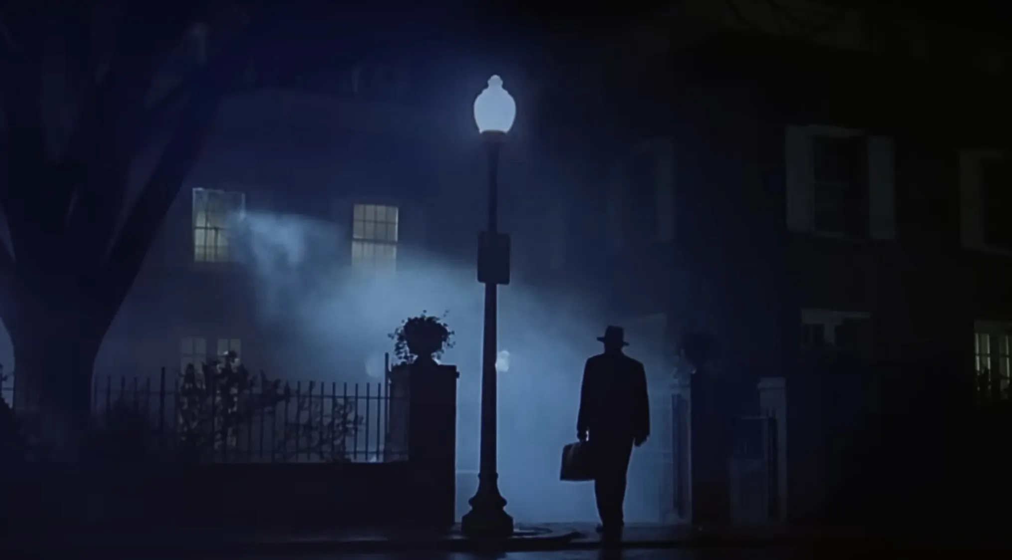 A lone figure in a fedora, carrying a briefcase, walks past a gas lamp post towards a house shrouded in mist. Horror movie trivia.