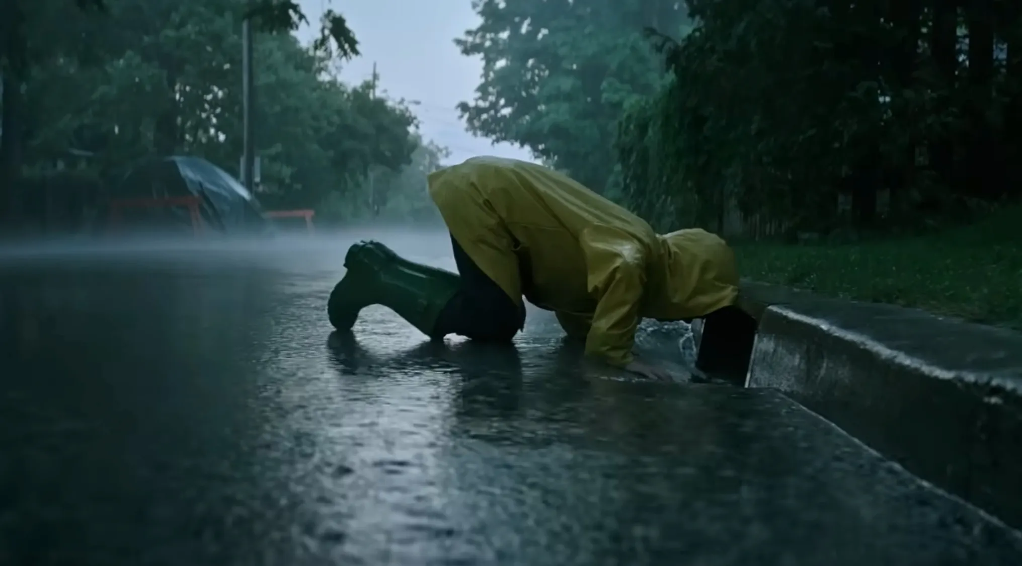 A child in a yellow raincoat and green boots reaches into a storm drain during a rainstorm. Horror movie trivia.