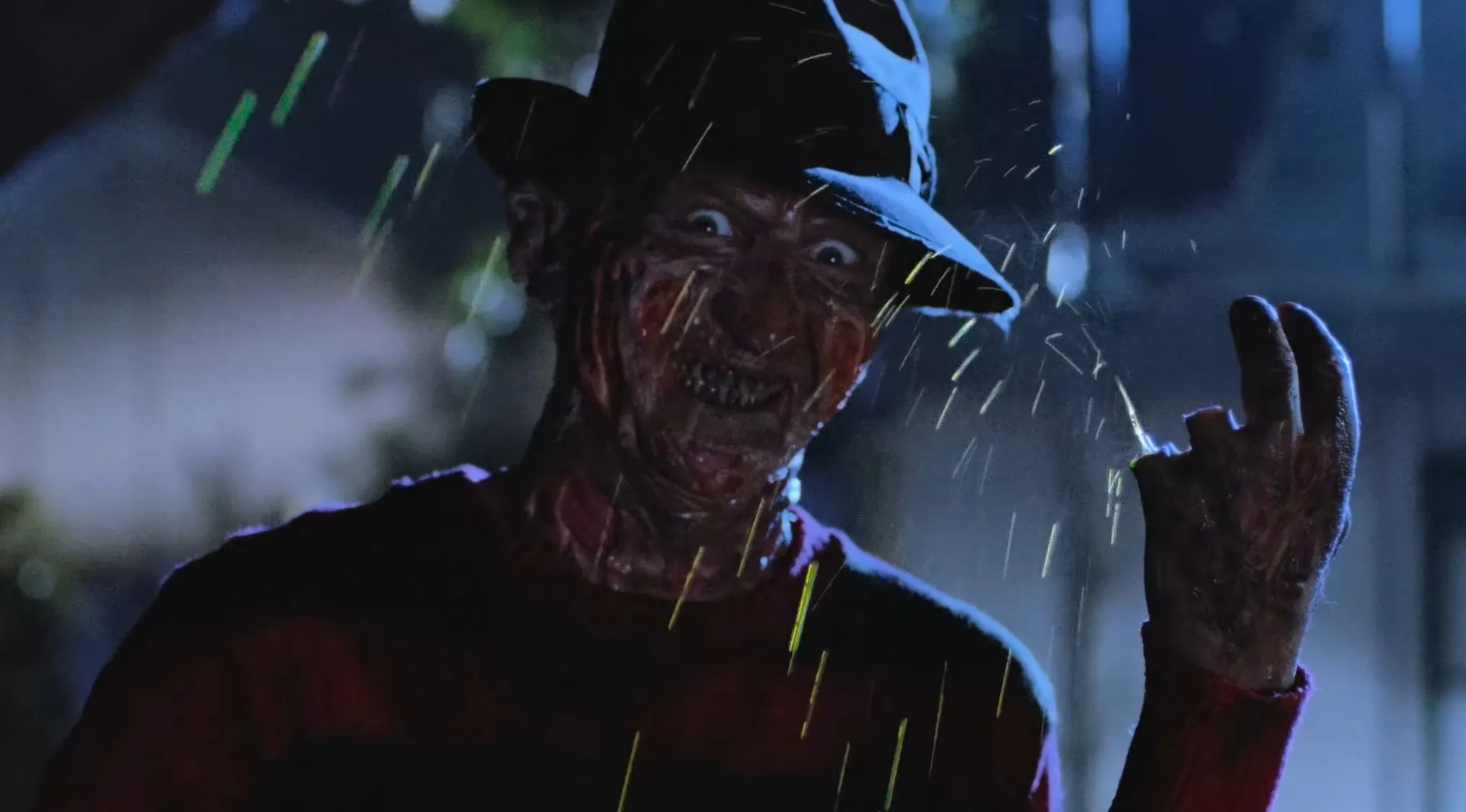 A close-up of Freddy Krueger in a hat and striped sweater, looking menacingly as rain falls. Horror movie trivia antagonist.