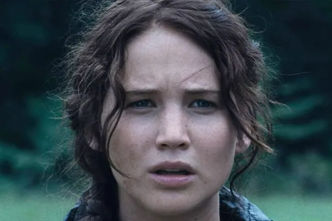 A close-up of Katniss Everdeen with a serious expression and braided brown hair, set against a blurred green background.