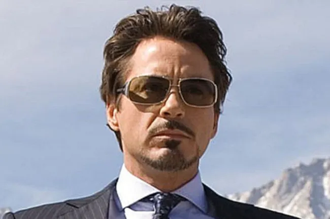 Tony Stark wearing sunglasses and a suit, with a serious expression against a mountain backdrop.