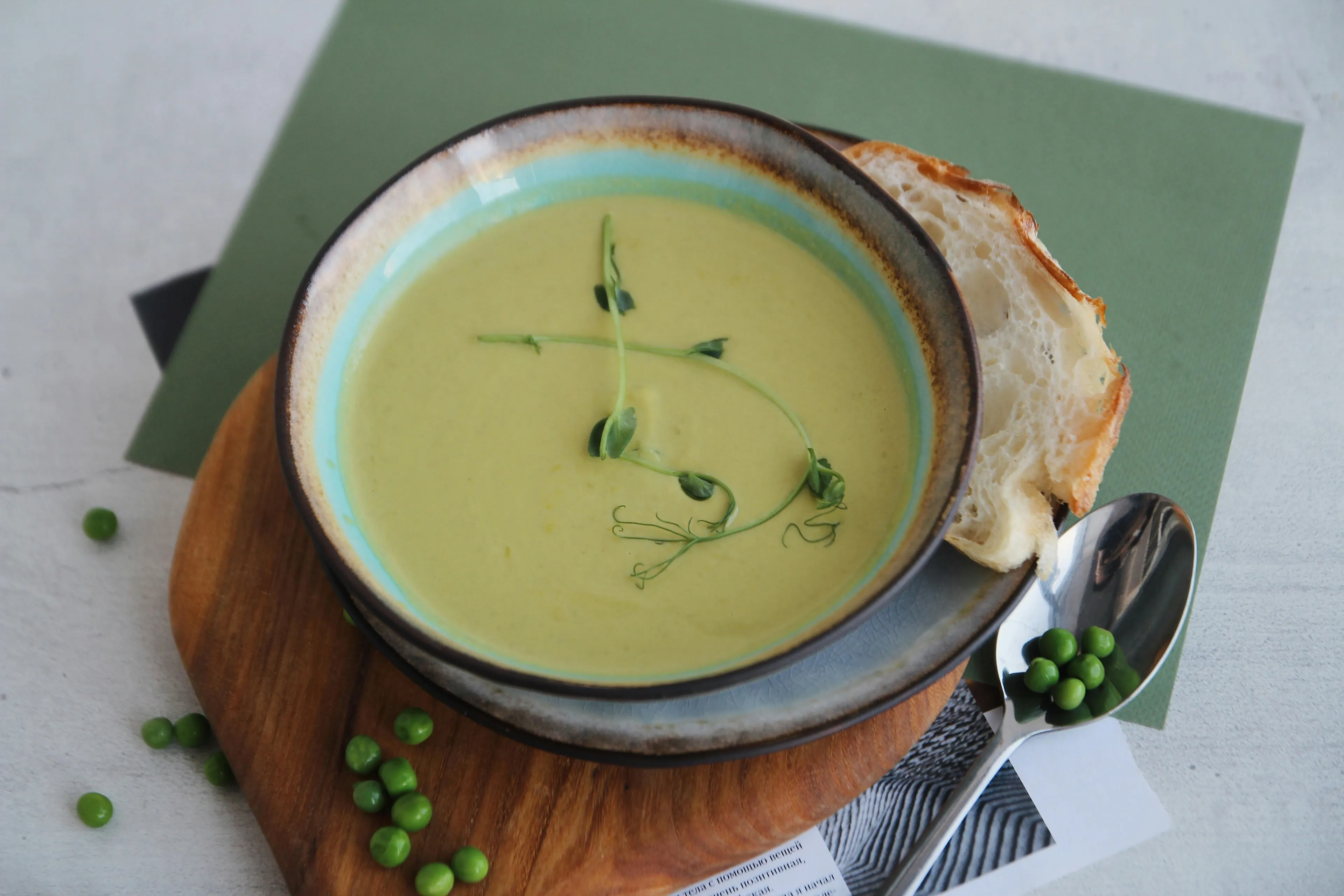 A cozy bowl of warm pea soup, garnished with pea shoots, served with a slice of bread and scattered peas.