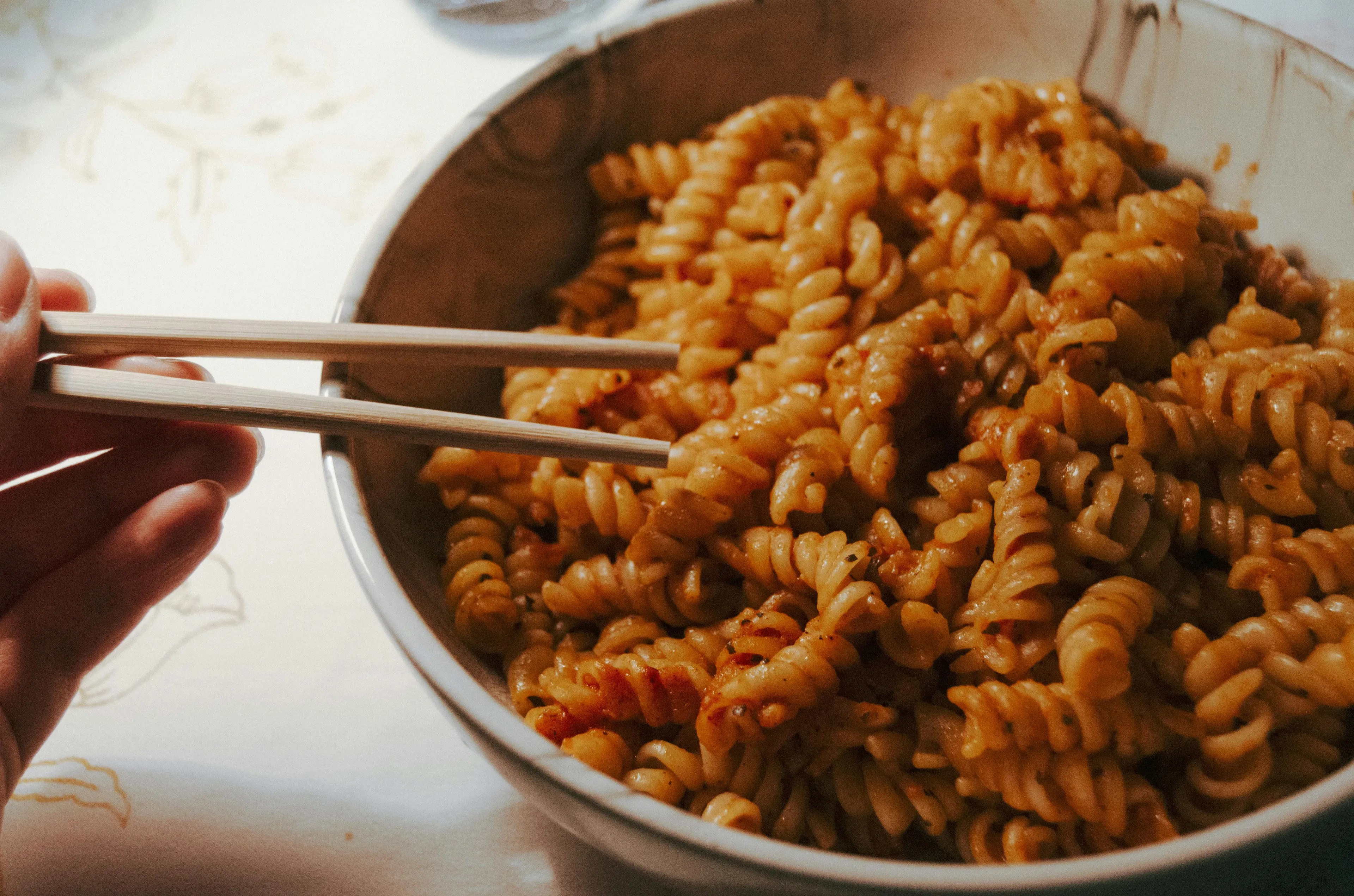 A hand holding chopsticks over a bowl of leftover pasta. The comfort food is ready to be enjoyed.